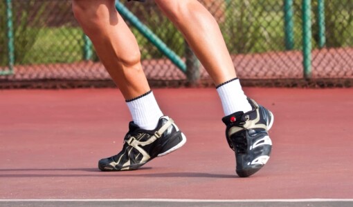 A person in black sneakers stands on a tennis court and flexes his calf muscles