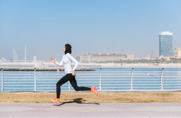 A woman wearing a black hijab runs on a paved, outdoor running path along the river, with a desert city skyline in the distant background.